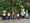 Ein Gruppenfoto zeigt eine fröhliche Gruppe von Menschen, überwiegend in Rollstühlen sitzend, vor einem Wegweiser mit der Aufschrift „Zu den Parkplätzen“. Zwei stehende Personen begleiten sie. Im Hintergrund sind Bäume und Parkautomaten zu sehen.