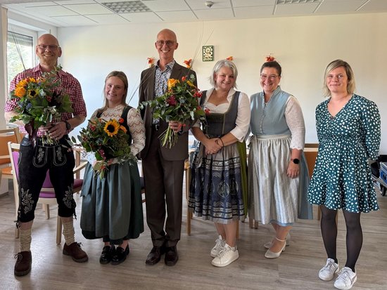 Sechs Personen stehen nebeneinander und lächeln. Vier halten Blumensträuße mit Sonnenblumen. Ein Mann und drei Frauen tragen traditionelle Tracht wie Lederhosen und Dirndls. Die anderen Personen tragen zivile Kleidung.