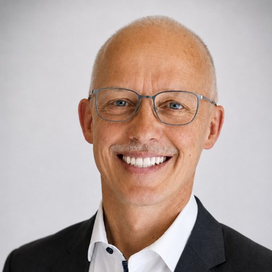 Günter Daubenmerkl Ein lächelnder Mann mit kahlgeschorenem Kopf und Brille trägt einen dunklen Anzug mit weißem Hemd. Er blickt direkt in die Kamera vor einem hellgrauen Hintergrund.