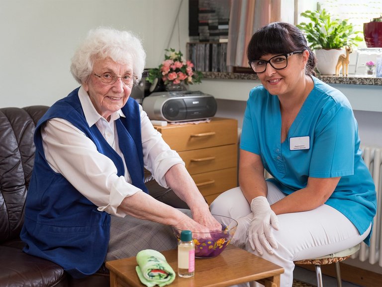 Eine Seniorin taucht ihre Hände in eine Schüssel mit lila Brei. Eine Pflegerin ist dabei