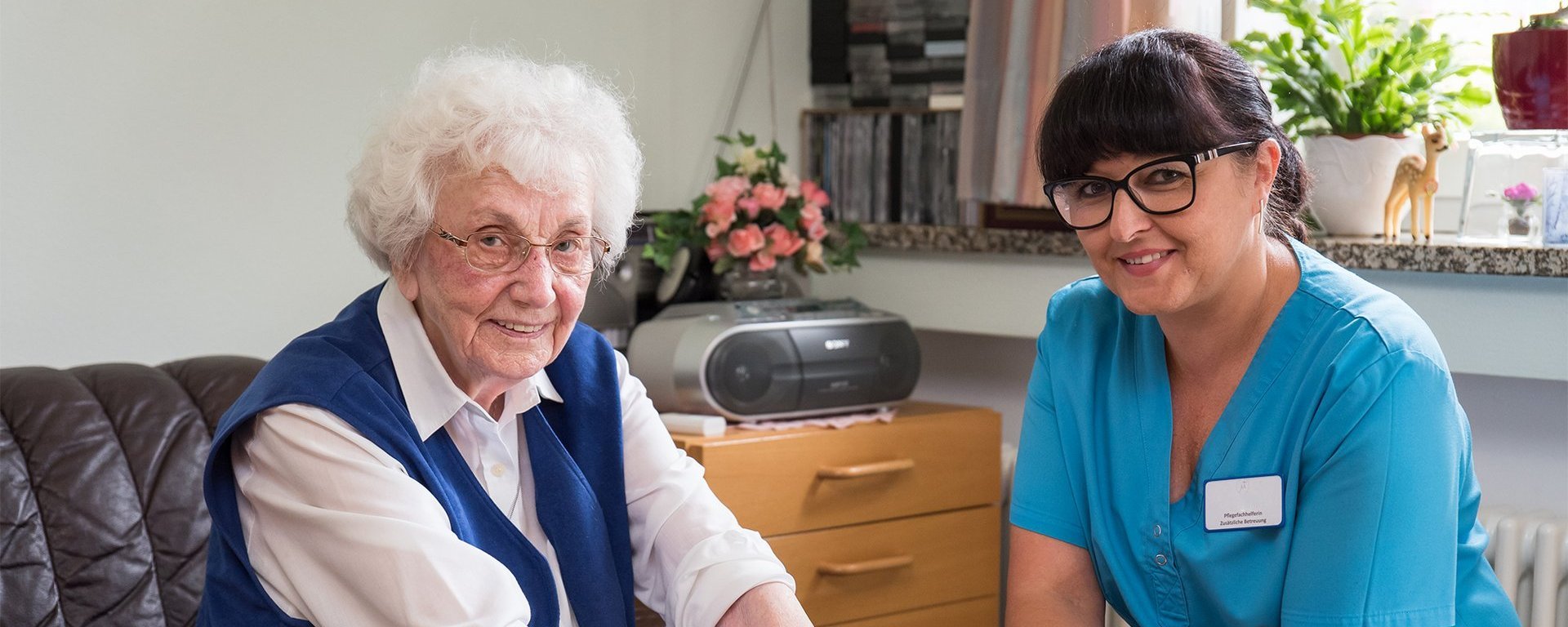 Eine Seniorin taucht ihre Hände in eine Schüssel mit lila Brei. Eine Pflegerin ist dabei